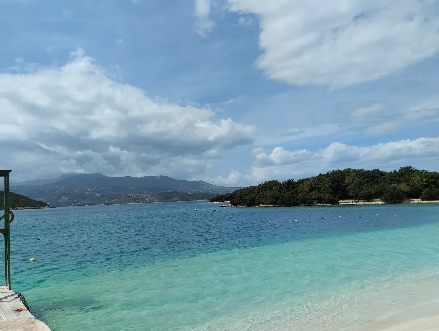 The original photograph: Ksamil beach in Albania, turquoise shallows, a wooded islet and a wooden jetty under a blue sky